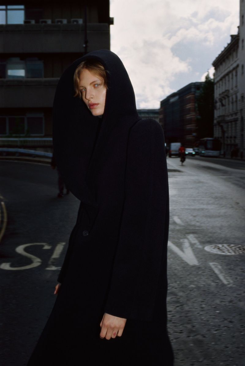 A female fashion model with fair skin wearing a black hooded coat on a city street with buildings and traffic in the background.