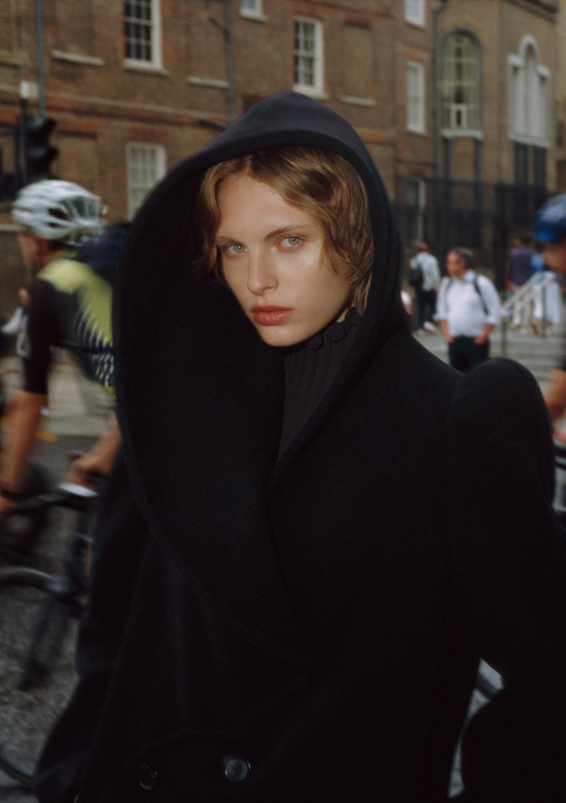 Fashionable young adult with blonde hair wearing a black coat with a hood standing on a city street with blurred cyclists.