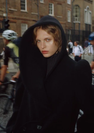 Fashionable young adult with blonde hair wearing a black coat with a hood standing on a city street with blurred cyclists.