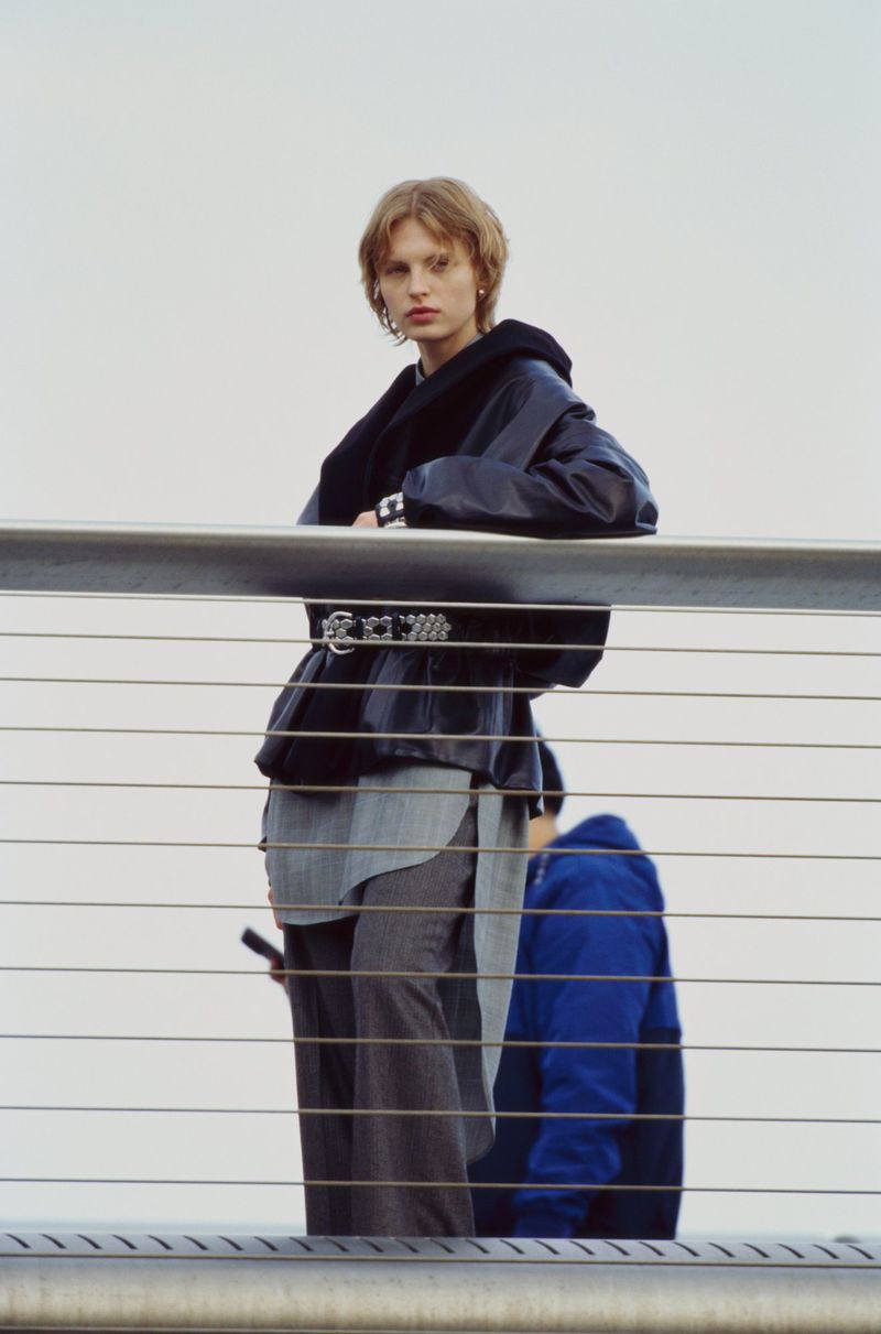 Fashion shot of a blonde adult female wearing a dark jacket and grey pants, posing near a metal railing with a grey sky background.
