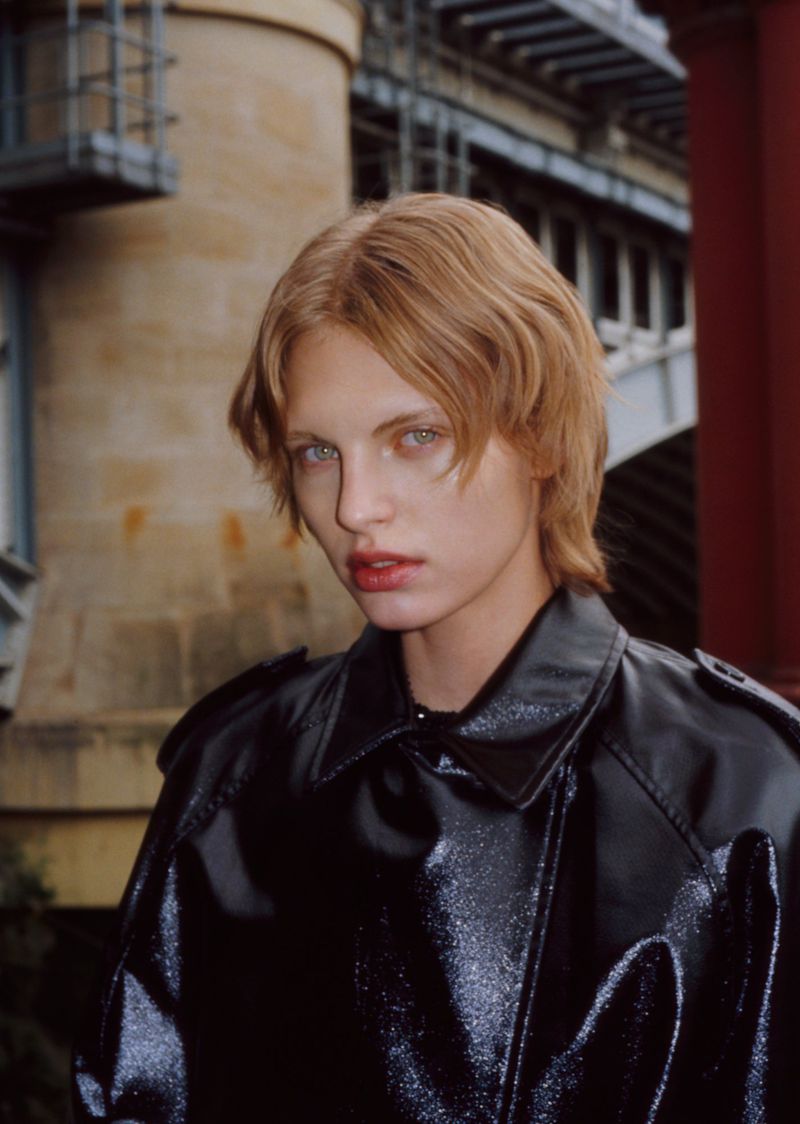 Close-up portrait of a blonde woman in a shiny black jacket against a gritty urban background with an architectural structure.