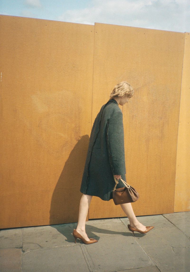 Stylish adult female walking outdoors in a coat and heels, carrying a brown handbag against a yellow wall and blue sky.