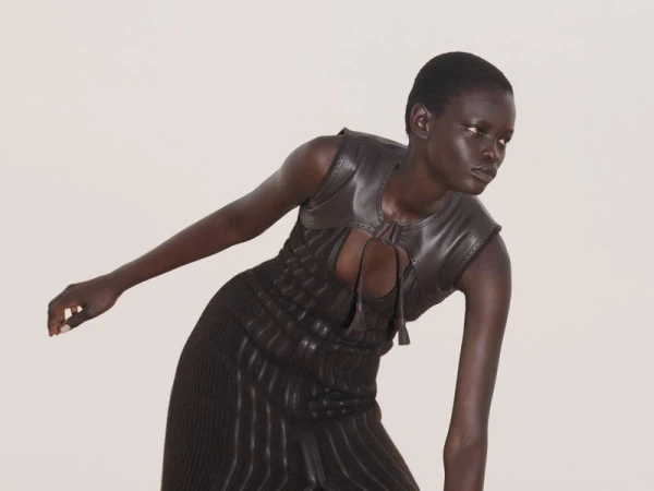 Woman in brown leather vest and knit dress poses in a studio with a neutral background for a fashion editorial shot.