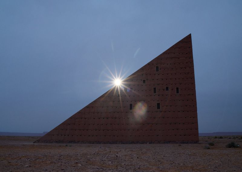 Architectural triangular building in a desert landscape with a bright sun starburst on its surface, under a blue sky with scattered clouds.