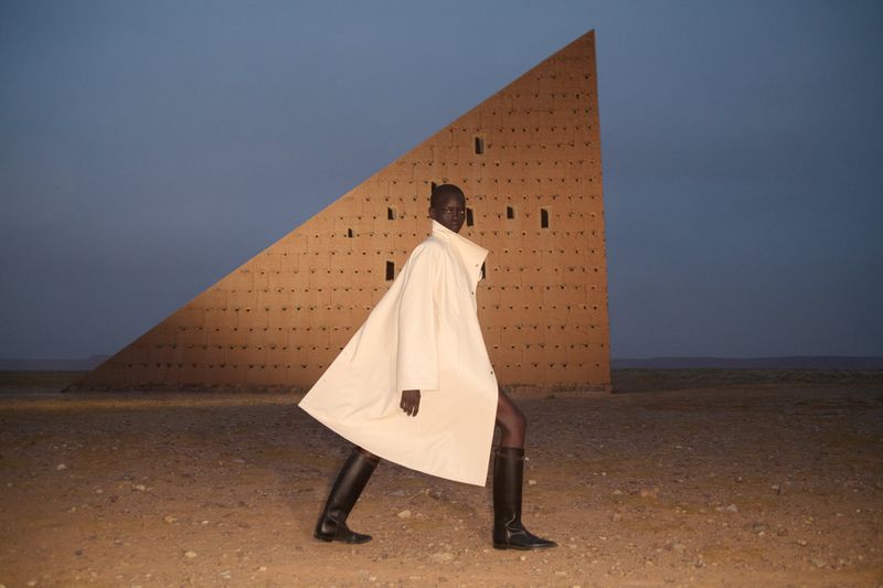 Fashion shot of a Black woman in a white coat and boots walking in a desert setting, with a geometric brown building behind her.