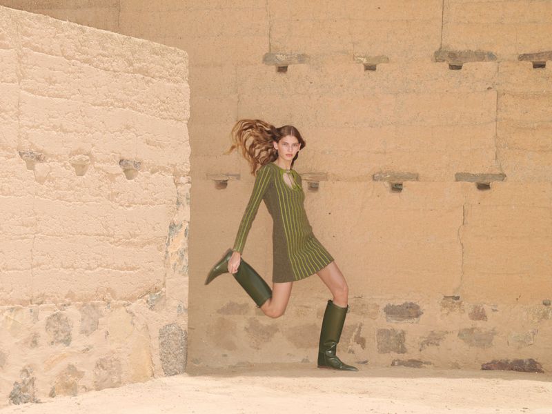 A fashionable woman wearing a green dress and boots poses mid-stride in a sunlit, architectural stone environment with earth-toned walls.