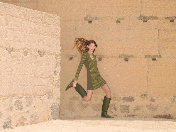 A fashionable woman wearing a green dress and boots poses mid-stride in a sunlit, architectural stone environment with earth-toned walls.