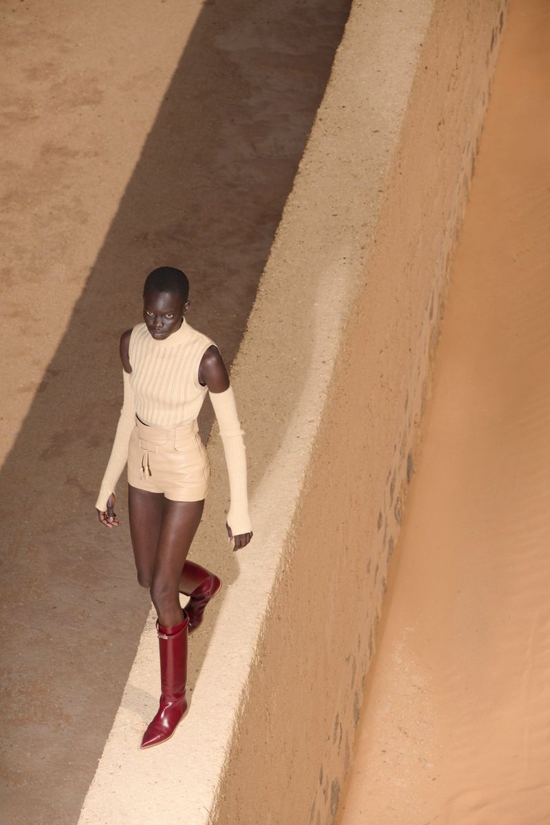A fashion model with dark skin is walking outdoors, wearing a cream-colored top and shorts, accessorized with red leather boots.