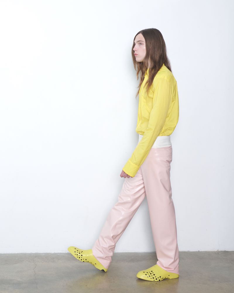 A person in yellow shirt and pink pants walks in yellow perforated shoes against a white wall and light grey floor in studio.