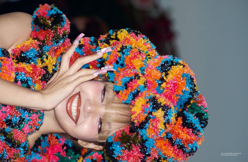 A blonde-haired female smiles with a multicolored pompom dress and manicured nails, indoor studio setting, vibrant colors, beauty shot.