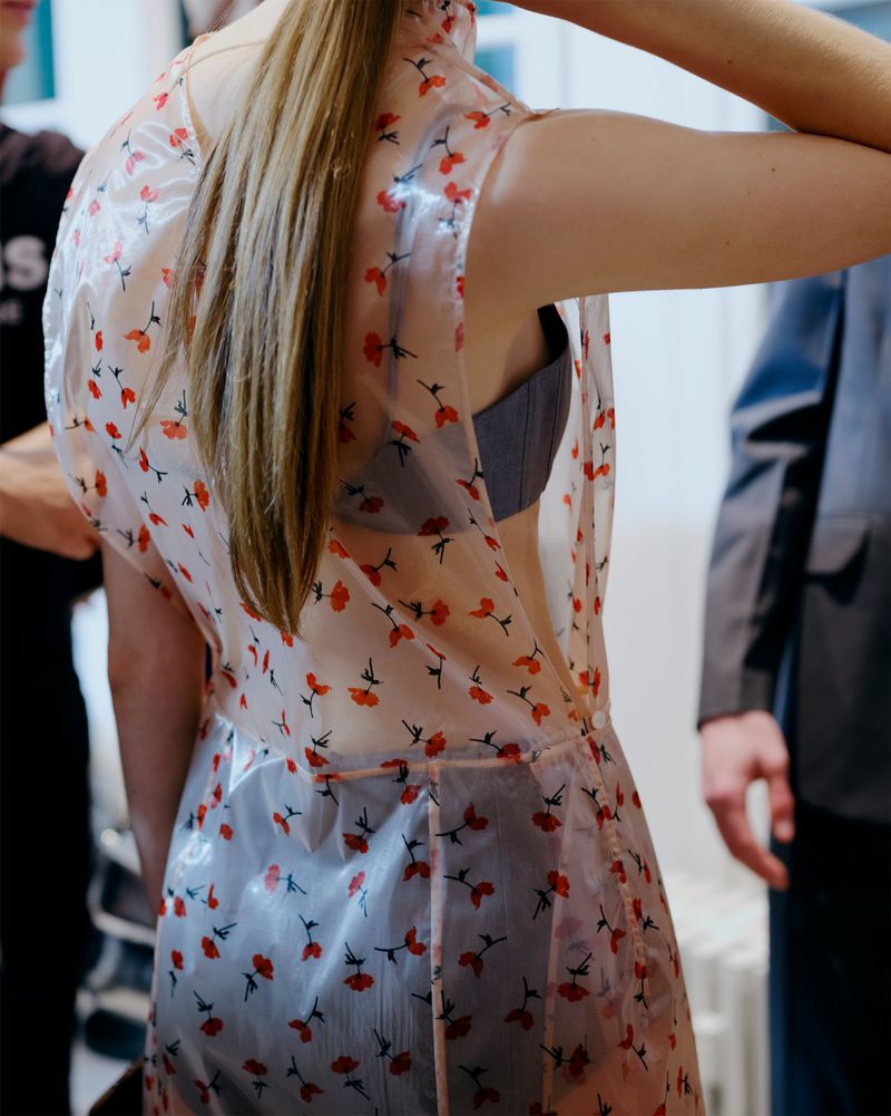 Back view of adult female with a transparent floral-patterned top, standing indoors with blurred figures in the background.