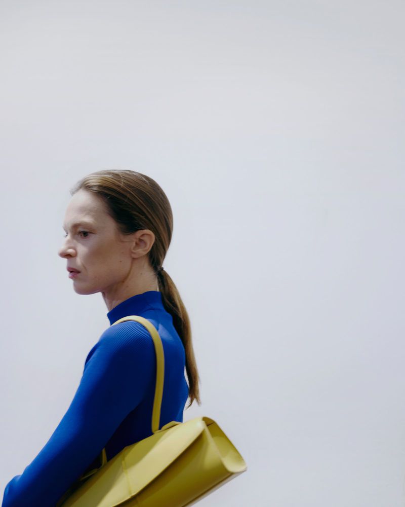 Woman with ponytail and blue long-sleeved shirt carrying a yellow bag, posing in front of a bright white background in a studio setting.