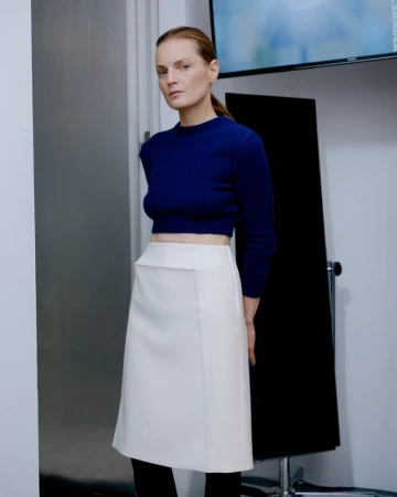 Woman in a blue long-sleeved crop top and white skirt posing in front of a grey wall, with a television in the background.