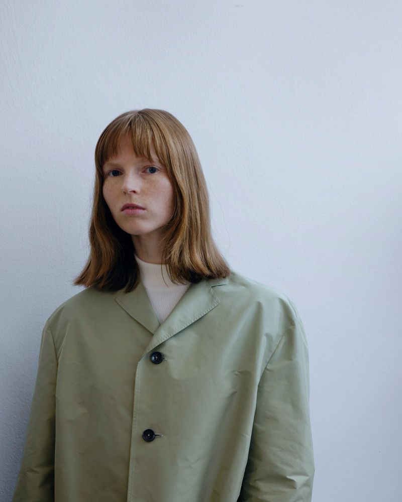 Model with shoulder-length hair wearing an oversized pale sage green buttoned jacket against a clean off-white studio background.