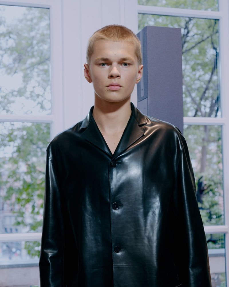 Portrait of a young adult male with blond hair wearing a black leather jacket indoors in front of a window.