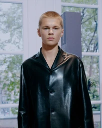 Model wearing a black leather buttoned collared jacket in front of large window panes looking outside.
