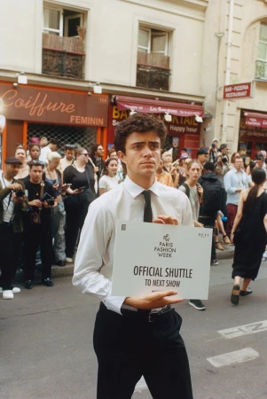 Fashion week attendee holds 'Official Shuttle' sign on street in Paris, surrounded by crowd and photographers, under daylight.