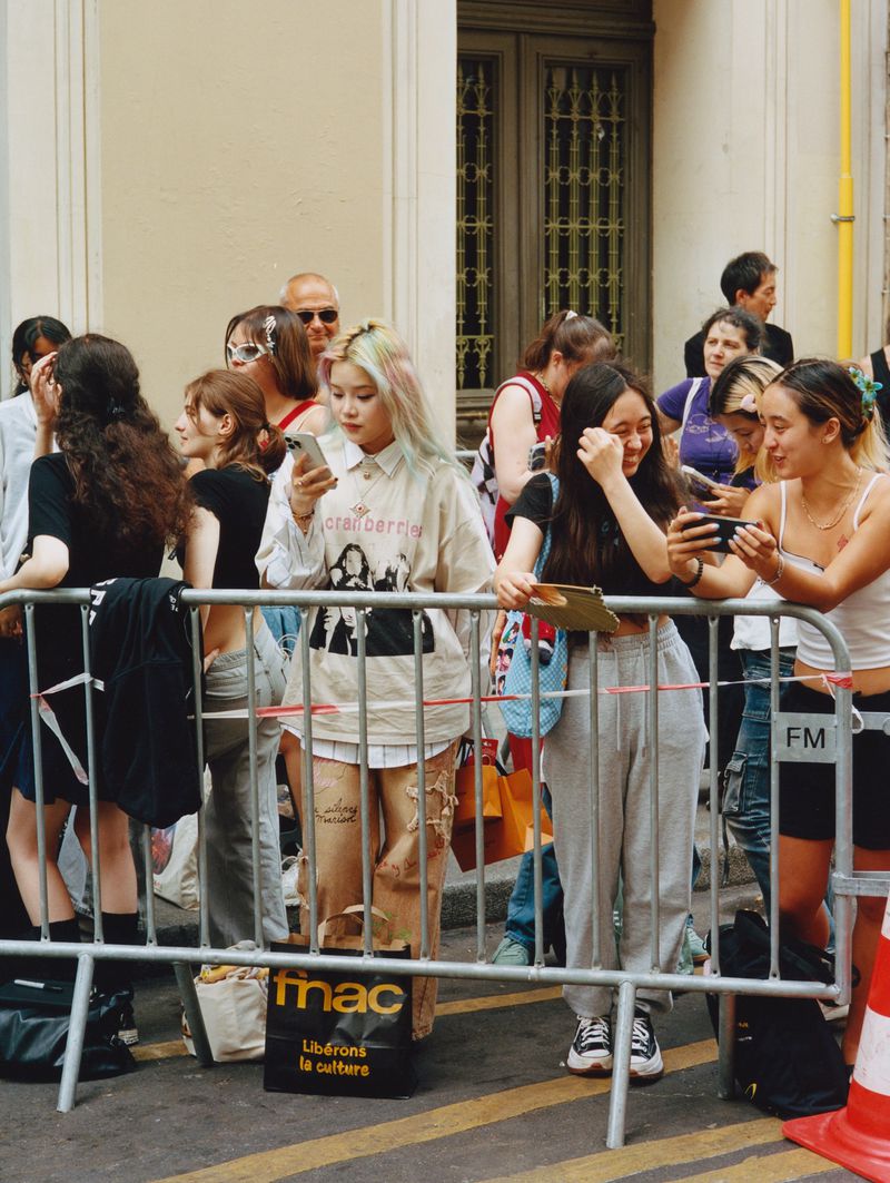 Outdoor daytime scene with a diverse group of people standing behind a metal barrier on a city street, some using phones and cameras.