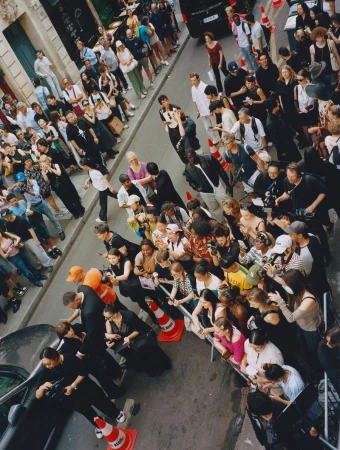 Crowd of people gather on a city street behind metal barriers, some holding cameras, under bright daylight, high angle view from above.