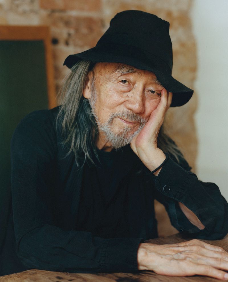 Older adult man with long gray hair, wearing black hat and jacket, resting his face on his hand, seated indoors at a table.