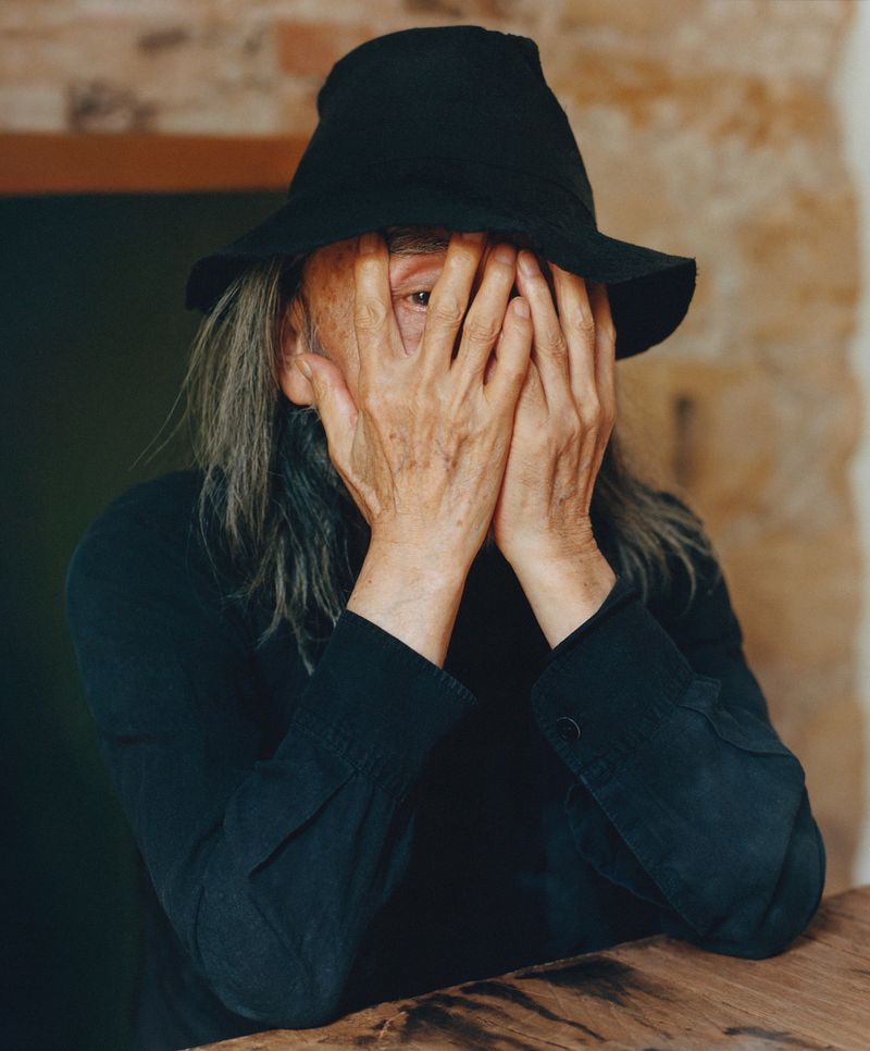 Portrait of an adult with hands covering the face, wearing a black hat and shirt, indoors against a neutral mottled background.