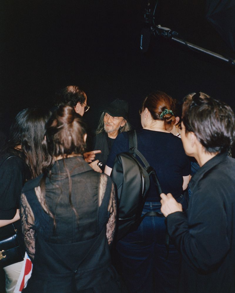 A diverse group of people surrounds an individual in a black hat, captured indoors with low lighting and visible film equipment overhead.