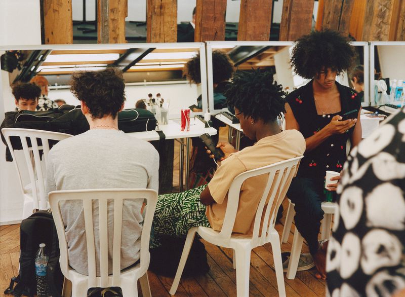 Several adult males are indoors in a room with wooden walls, looking at their mobile phones while seated in white chairs at tables.