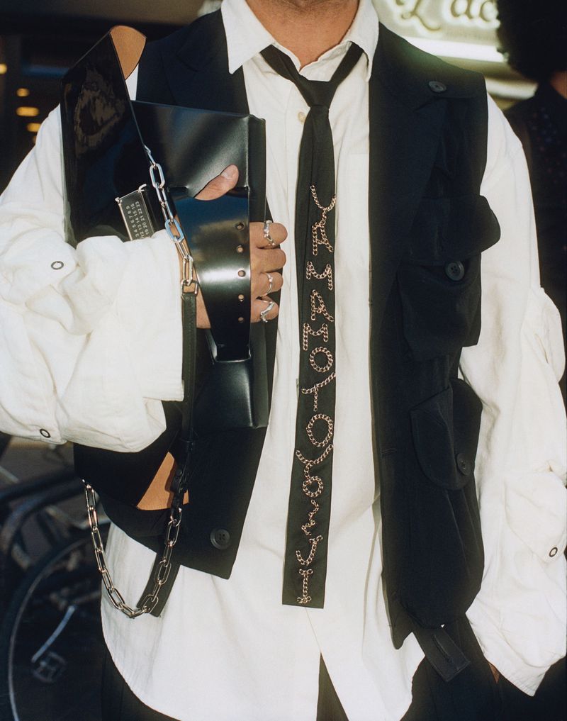 Fashion shot featuring a person in a white shirt and black vest with chain accents, holding a black clutch, in an outdoor urban setting.