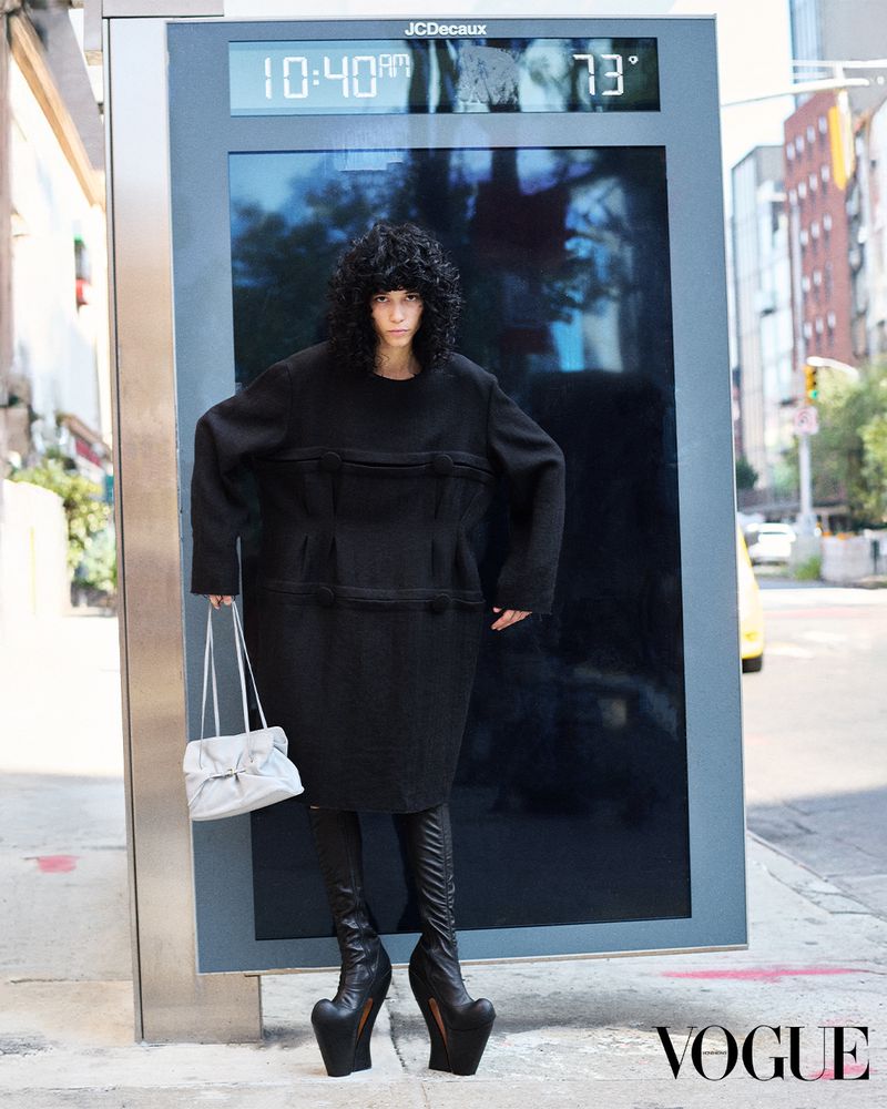 Woman in oversized black dress and sculptural high heels stands by digital display with time, temperature in city street setting.