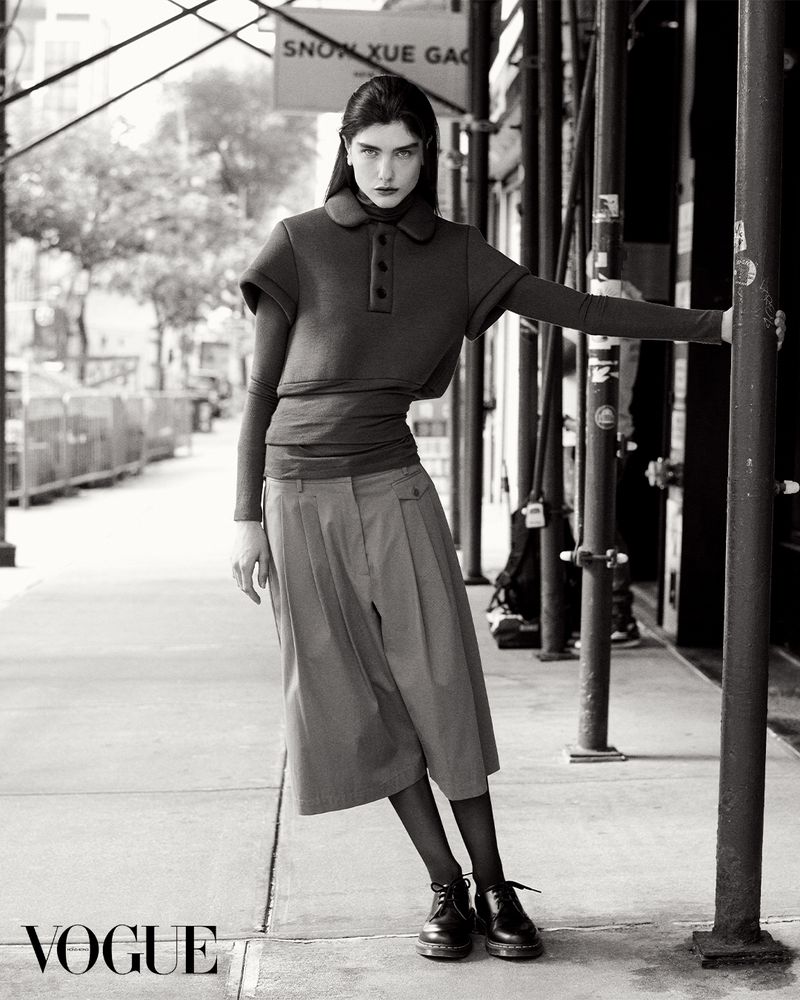 A brunette woman in a dark top and khaki culottes poses in front of a building with scaffolding on a city sidewalk in black and white.