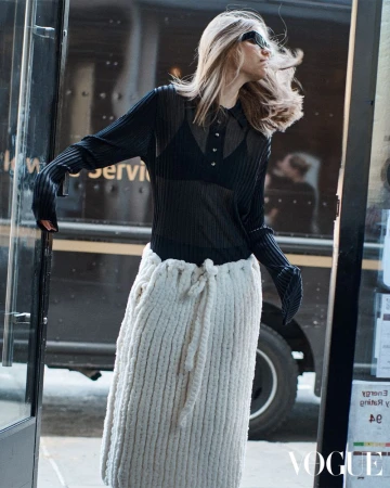 Fashion editorial shot: a female model in a black sheer top and long knit skirt poses near a dark service vehicle in natural light.
