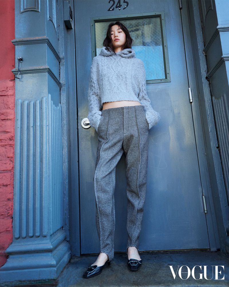 A young Asian woman stands in front of a blue door wearing a grey sweater, herringbone pants and black patent leather loafers.