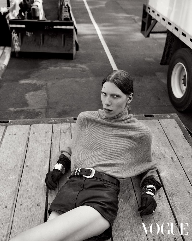 Black-and-white fashion shot of a stylish woman in a poncho and shorts lying on a wooden surface near heavy equipment and trucks.