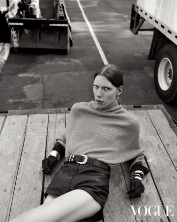 Black-and-white fashion shot of a stylish woman in a poncho and shorts lying on a wooden surface near heavy equipment and trucks.