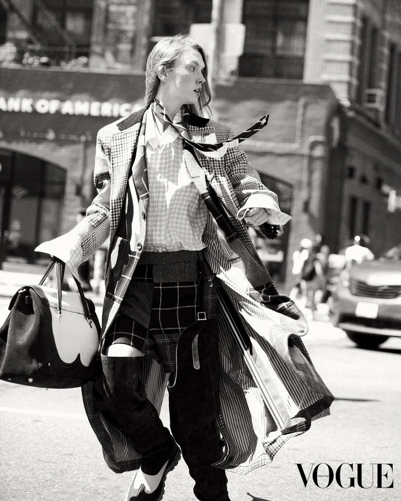 Fashionable person in patterned coat and pants walks across a city street holding a bag in black and white Vogue editorial.