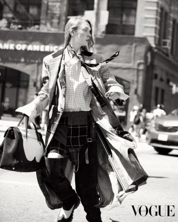 Fashionable person in patterned coat and pants walks across a city street holding a bag in black and white Vogue editorial.