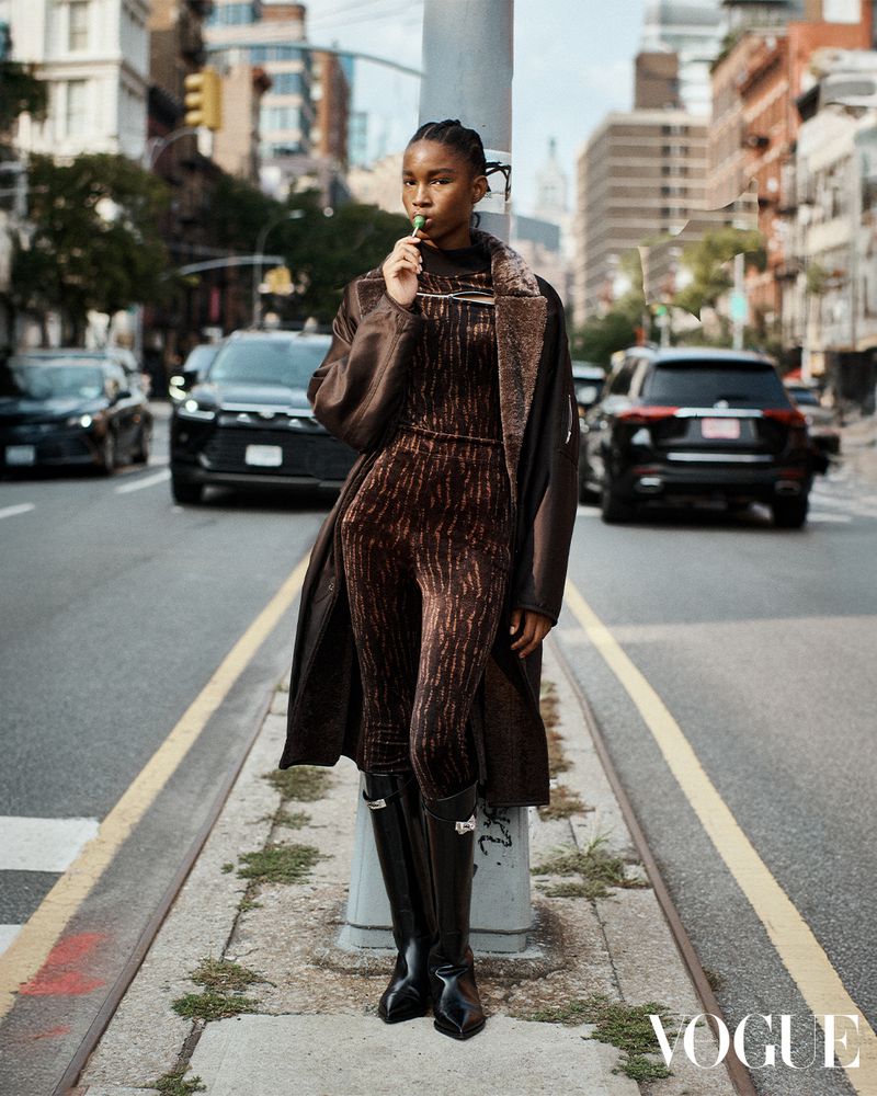 A fashionable young woman in a brown jumpsuit and long coat poses in the street eating a lollipop with cars and buildings in the background.
