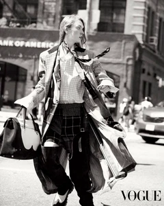 Black and white photo of a fashion model walking across a city street carrying a large two-tone leather handbag in a layered patterned coat.