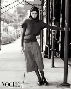 Model in dark layered top and skirt leaning against a scaffolding pole on a city sidewalk in a black and white fashion editorial image.