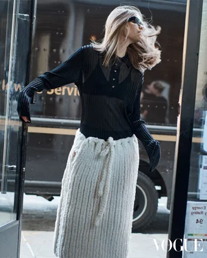 Model wearing dark sunglasses, a sheer black top, and a chunky cream knit maxi skirt stepping through a glass doorway.