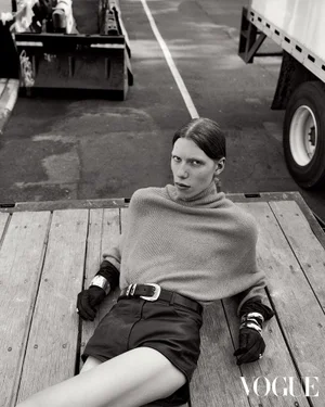 Model reclining on a wooden truck bed wearing a turtleneck knit sweater, leather shorts, and black gloves in black and white.