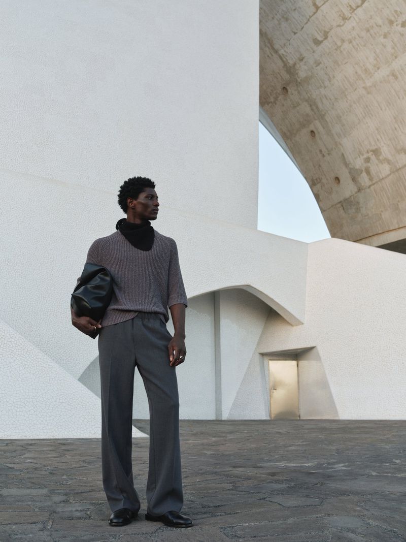 An adult male model posing outdoors in gray knit sweater, black turtleneck and gray pants with a black leather bag under his arm.