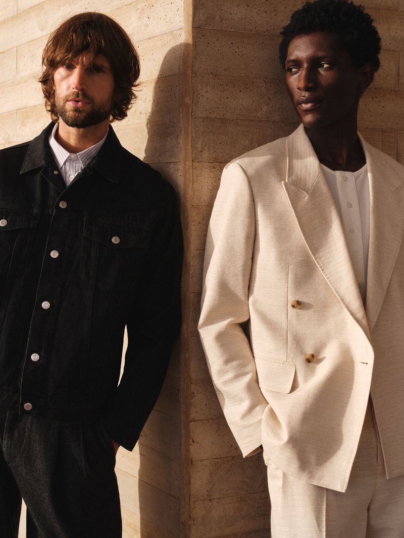 Two men in fashionable jackets pose near a textured wall. One wears a black denim jacket, the other a cream-colored linen suit jacket.
