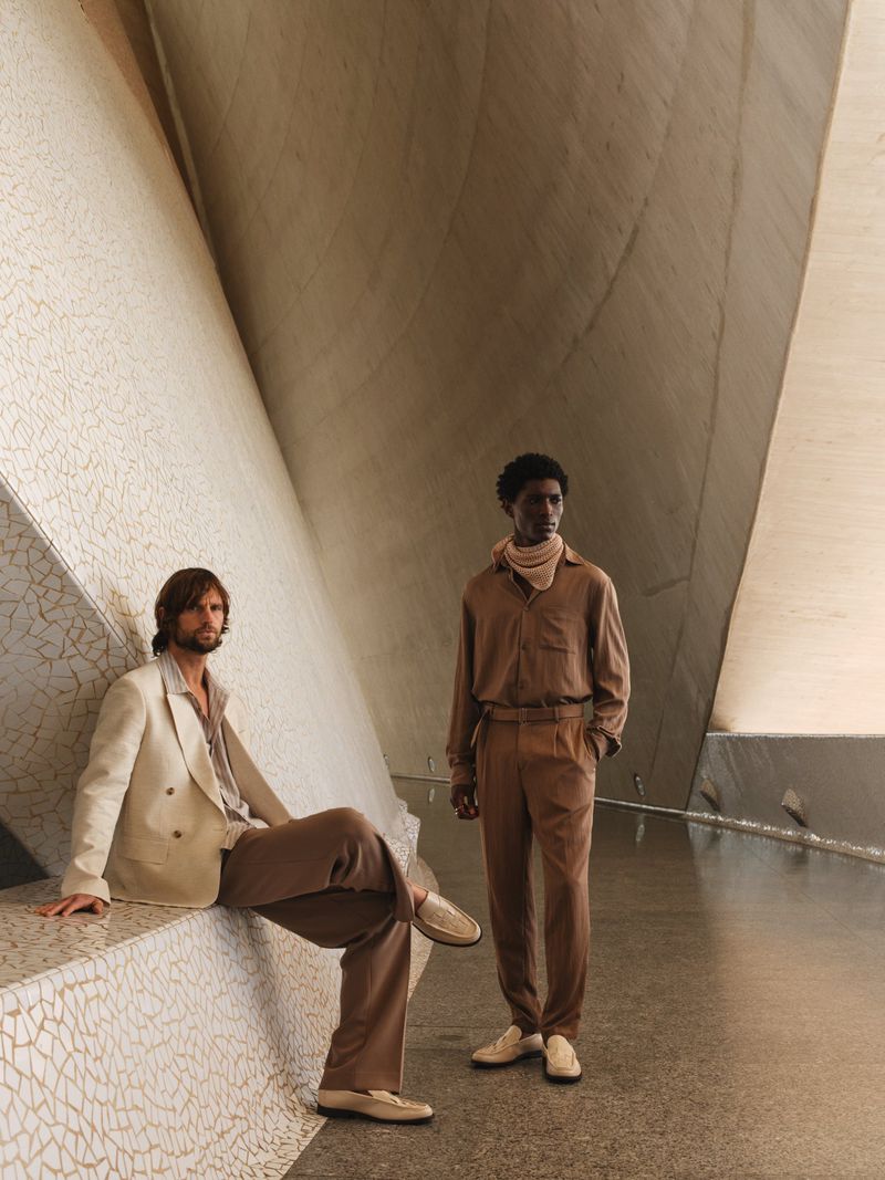 Two adult males pose indoors in a modern space with neutral colors. One sits in a beige suit, the other stands in brown. 