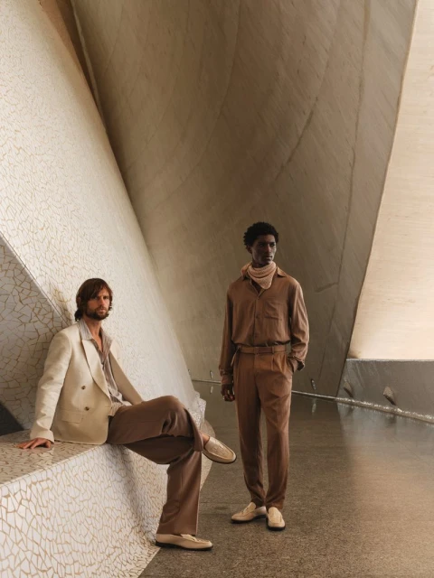 Two adult males pose indoors in a modern space with neutral colors. One sits in a beige suit, the other stands in brown. 