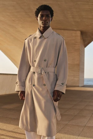 Model in a long beige belted trench coat standing outdoors against architectural concrete structures.