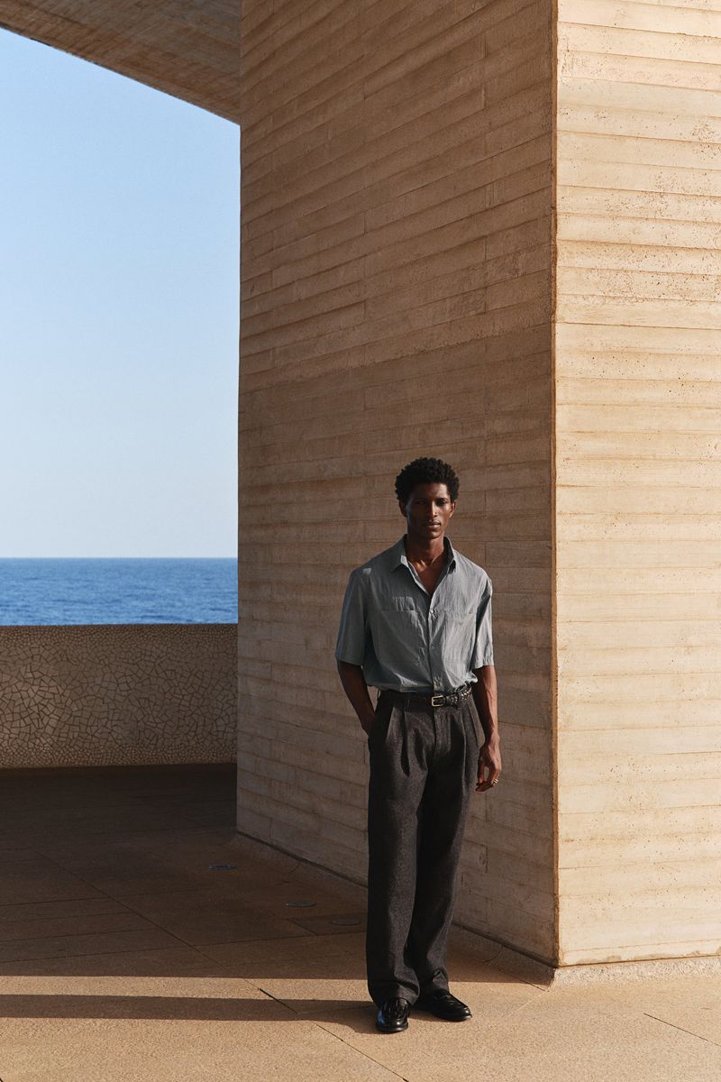 A lone male model stands on a stone terrace overlooking the ocean wearing a light gray button-down shirt and charcoal wool slacks.