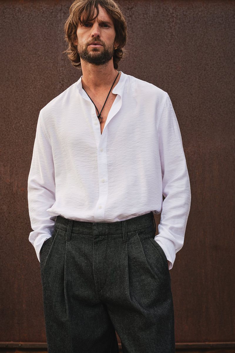 A stylish adult male with a beard stands outdoors, wearing a white linen shirt and gray pleated trousers against a neutral background.