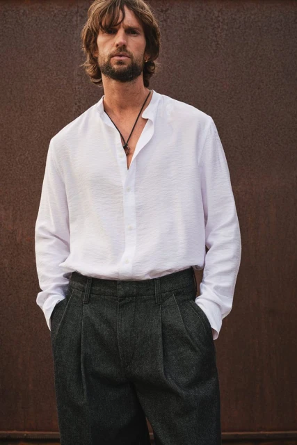 A stylish adult male with a beard stands outdoors, wearing a white linen shirt and gray pleated trousers against a neutral background.