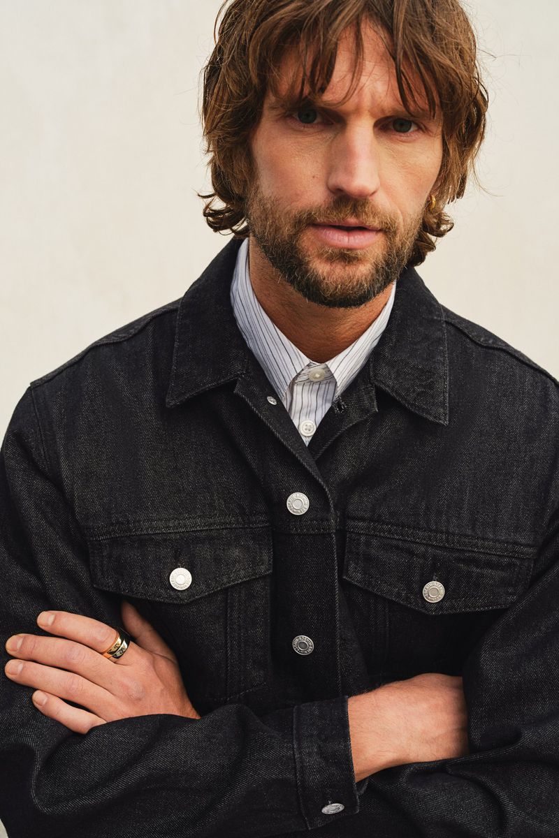 Portrait of a man with medium brown hair, wearing a black denim jacket over a light blue striped shirt, against an off-white backdrop.